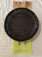 Top view of a brown woven round Japanese basket showing triangular weave pattern and accompanying artist name labels with Japanese text.