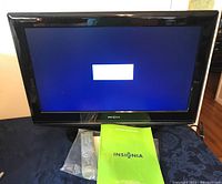 Front view of the 26-inch Insignia LCD TV turned on with remote control and instruction manual placed in front of it.