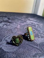 Two rings placed on a dark fabric background, one heart-shaped with a green stone, the other rectangular with green and blue stones and small blue bead accents on the sides.