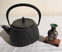 Black cast iron mini teapot with handle and spout beside a mini blue ceramic vase on a wooden stand.