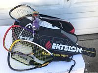 Photo showing three racquetball racquets with two protective cases and a canister of purple racquetballs on top of the Ektelon bag.