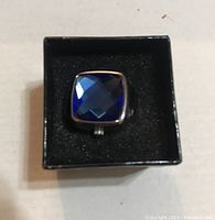 Top-down view showing the sapphire-colored faceted stone set in square silver-tone casing placed in black foam inside a box.