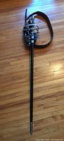 Silver rapier sword sheathed in black belt holder with multiple straps displayed on hardwood floor.