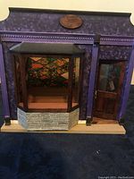 Front view of the unfinished dollhouse dining room showing purple exterior, bay window with stone base, door with window, and colorful interior wallpaper.