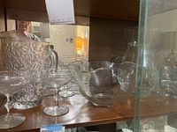 Glass pitchers, bowls, and clear glassware displayed on a shelf with a reflection of a person in a mirror.