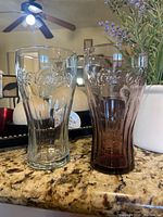 Two Coca Cola glasses on granite countertop, one clear and one tinted with embossed Coca Cola logo.