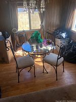 Photo showing the full dinette set with a square glass top dining table and four wrought iron chairs with beige cushions.