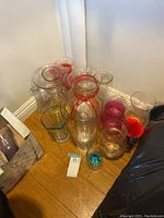 Photo showing multiple glass vases of various shapes, some tied with red ribbons, and some containing colored glass stones on a wooden floor corner.