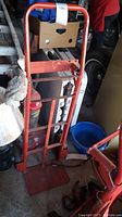 Full view of the red metal hand truck standing upright, showing the handle, frame, and base plate with visible scratches.