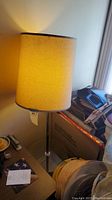 Floor lamp with yellowish fabric lampshade, standing beside some household items and boxes, providing full view of the lamp height and lampshade texture.
