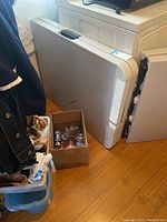 Folded plastic rectangular table leaning against laundry machine with cardboard box nearby, showing size and overall form.