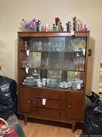 Front view of Mid-Century Modern wooden china cabinet filled with glassware and dishes inside glass shelves. Two drawers and two cabinets below.