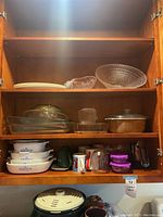 Wooden kitchen cabinet with shelves containing Pyrex glass baking dishes, Corning Ware baking dishes with blue cornflower pattern, clear textured glass serving bowl, plastic divided plates, coffee cups and mugs, and plastic storage containers.