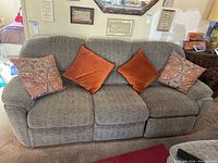 Front view of three-seat gray fabric recliner couch with four throw pillows placed on it in a well-lit living room.