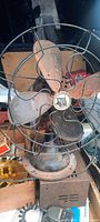 Front view of vintage 1930's metal fan showing four rusted blades behind protective cage and emblem at center