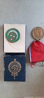 Photo of two antique jacket badges, one green enamel badge in original blue box, and one bronze medal with red, white, and blue ribbon.