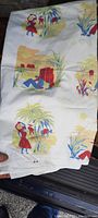Full view of the folded and unfolded tablecover showing colorful vintage pattern with people, reeds, and nature elements on white fabric.