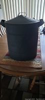 Photo of black painted metal steamer can, showing cylindrical shape with two side handles and fitted lid, placed on a wooden dining table.
