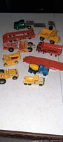 Photo shows multiple antique/vintage metal construction toys on a white surface. Green semi truck with trailer, various yellow construction vehicles, a blue and yellow cement mixer truck, red tanker truck, and red metal structure are visible.