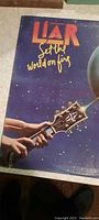 Front cover of the Liar LP record showing the title 'Set the World on Fire' with a hand playing a guitar and a glowing globe in space