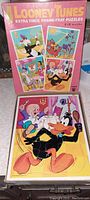Photo showing front of original pink box with four Looney Tunes puzzle illustrations and one assembled puzzle featuring Daffy Duck and Elmer Fudd on a dinner table