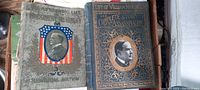 Two hardcover antique books about President William McKinley side by side in a box. One book has a gray-green cover with an American flag shield and portrait; the other has a dark ornate gold embossed cover with a photo portrait and title.