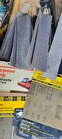 Several stacks of gray belt sander strips and unopened packs of sandpaper sheets with visible brand and grit info.