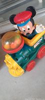 Front angled view showing Mickey Mouse figure wearing a red hat, on a green and yellow toy train with red wheels and a transparent dome with orange balls in front.