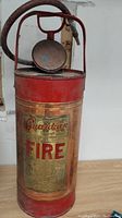 Front view of antique fire extinguisher in red and copper with 'Guardian FIRE' label plaque on body, featuring pressure gauge and hose on top with handle.