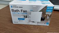 Front view of boxed DewStop Breeze Easy bath fan with fan control, showing product name and features including humidity sensing fan control and quiet operation.