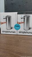 Two unopened Simplehuman touch-free sensor pump soap dispensers in original packaging, showing front with hand triggering soap dispensing.