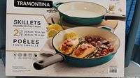 Front view of the Tramontina box showing the 2-piece enameled cast iron skillet set, with cooking scene images of salmon and vegetables inside turquoise skillets.