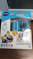 Front of sealed retail box showing product image of blue and white VTech KidiZoom PrintCam camera and main features including instant inkless printing, 5MP photo resolution, print up to 320 photos and 80 stickers, suitable for children 4+ years.