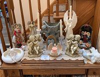 Lot of decorative angel figurines and candle holders arranged on wooden table with doily, exhibiting a variety of materials, colors, and themes including cherubs and Christmas angels.