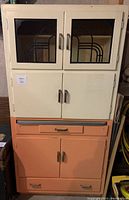 Front view of antique hutch showing upper white cabinet with glass doors and lower peach cabinets and drawers