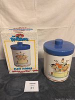 Ceramic cookie jar shown beside original packaging box on gray fabric surface with white backdrop.