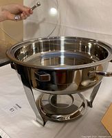 Shiny stainless steel round gourmet chafing dish with handles, elevated frame for heat source, shown open without lid.