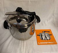 Pressure cooker with black handles and lid, positioned alongside an orange booklet showing Lagostina branding and cooker illustration.