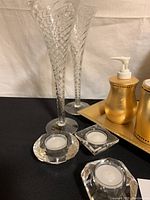 Two tall, twisted stem Romanian hand blown champagne flutes, two gold ceramic bath set items, three glass tealight holders with candles on a black cloth