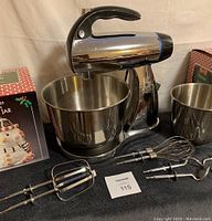 Sunbeam Mixmaster standing mixer with one stainless steel bowl inserted, one separate bowl and three types of metal mixing attachments displayed on black cloth.