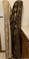 Two large fabric bolts leaning against a wall next to kitchen cabinets showing one gold brocade style fabric bolt and one burgundy floral brocade bolt.