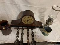Shows the wooden Ingraham mantle clock, glass lantern, MacDonalds tin, metal hanging utensils with decorative handles, and part of the Rogers pottery jug.