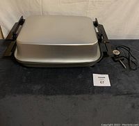 Electric skillet with rectangular metal lid, black handles on each side, and power cord with attached temperature control probe.