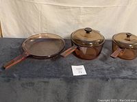 3-piece set of amber glass Visions Corning cookware arranged on fabric surface: 2 sauce pots with lids and 1 skillet.