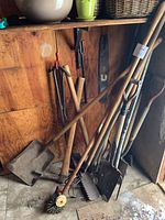 Full view of various gardening tools leaning against a wooden wall including shovels, pickaxe, hoes, and a spiked cultivator tool.