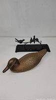Brass duck figure in front with brass birds perched on a branch sculpture on a black base in the background