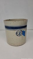 Front view of cream colored stoneware crock with two blue horizontal stripes near the top and circular Empire Pottery mark with number '2'.