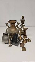 Group lot of various brass items arranged on a white surface including a large brass vase with dragon handles, brass candlesticks, metal pitcher, small brass vases, pyramids, and small decorative pieces.