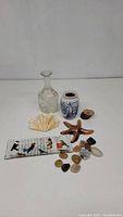 Photo showing group lot of decorative items including glass bottle decanter, porcelain vase, starfish figurine, glass bird tray, seashell cluster, and various decorative stones.