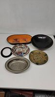 Front view of six assorted plates and trays on white surface showing variety of materials including resin, ceramic, silverplate, and wood/lacquer.